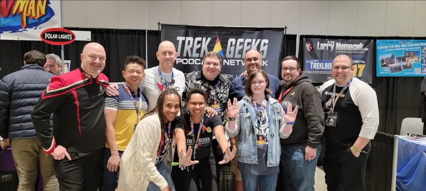 Ten Trek Geeks hosts crowded together in front of the network’s banner at a convention booth, several of them giving the Vulcan salute