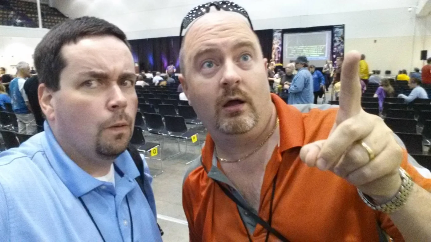 Bill Smith and Dan Davidson in a convention hall, mugging for a phone camera, Dan pointing off to one side